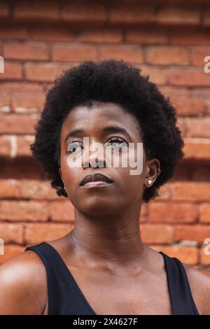 Young unhappy African American female with curly hair standing against brick wall and thoughtfully looking away Stock Photo