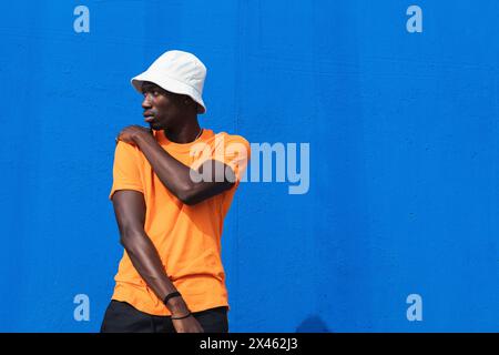 Young African American male in bright yellow t shirt and sportive pants posing while standing against blue wall Stock Photo