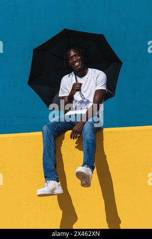 Low angle full body of optimistic young black male in casual outfit with panama hat and sunglasses smiling happily while holding an umbrella siting on Stock Photo