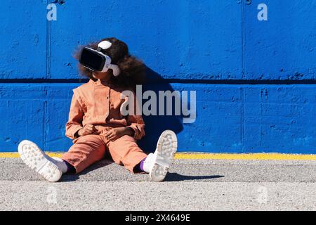 Unrecognizable African American girl in trendy suit and VR headset exploring virtual world while sitting against blue wall on the street Stock Photo