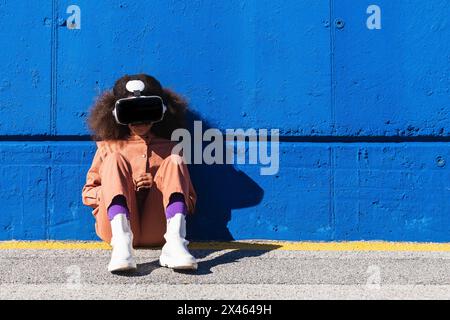 Unrecognizable African American girl in trendy suit and VR headset exploring virtual world while sitting against blue wall on the street Stock Photo