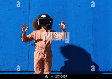 Unrecognizable African American girl in trendy suit and VR headset touching air while exploring virtual world against blue wall on street Stock Photo