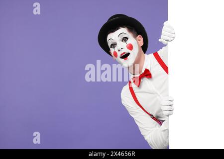 Funny mime artist peeking out of blank poster on purple background. Space for text Stock Photo