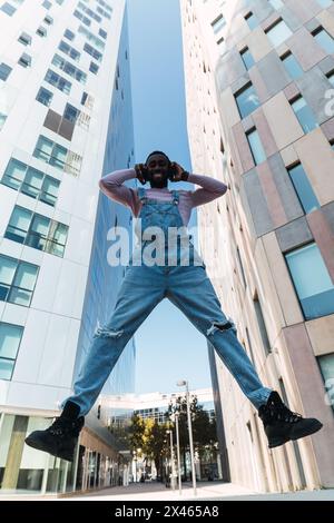 From below of young African American male in cool outfit jumping above ground on street while listening to songs in headphones and looking at camera Stock Photo