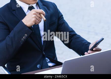 Confident Asian male entrepreneur sitting at table in cafe and smoking cigar while browsing smartphone Stock Photo