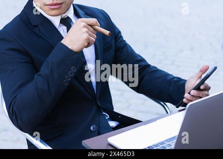 Confident Asian male entrepreneur sitting at table in cafe and smoking cigar while browsing smartphone Stock Photo