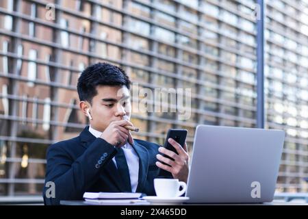 Confident Asian male entrepreneur sitting at table in cafe and smoking cigar while browsing smartphone Stock Photo
