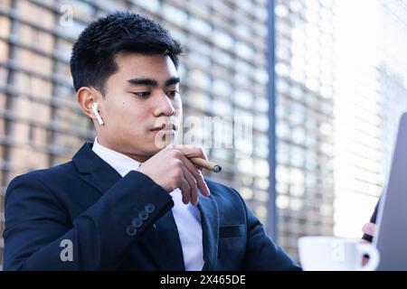 Confident Asian male entrepreneur sitting at table in cafe and smoking cigar while browsing smartphone Stock Photo