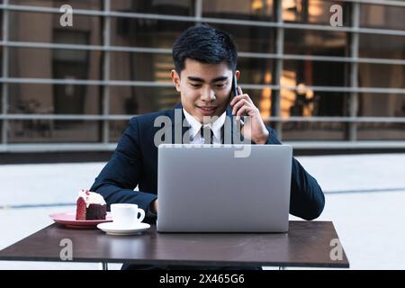 Smiling Asian male entrepreneur sitting at table in street cafe and working remotely while typing on laptop and speaking on smartphone Stock Photo