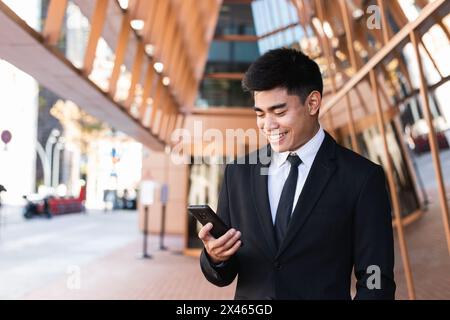 Delighted Asian businessman standing in street and surfing Internet on mobile phone Stock Photo