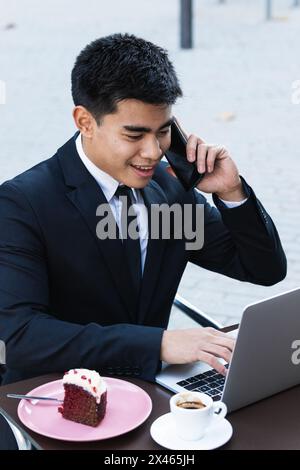 Smiling Asian male entrepreneur sitting at table in street cafe and working remotely while typing on laptop and speaking on smartphone Stock Photo