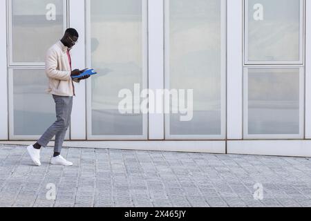 Side view of busy African American male freelancer in smart casual style walking along street and browsing mobile phone Stock Photo