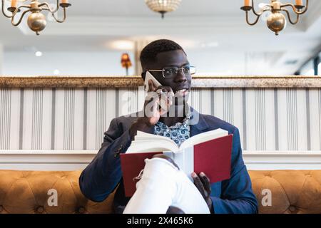 Young African American male executive with agenda talking on cellphone while sitting with crossed legs on couch and looking away Stock Photo