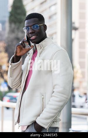 Young content African American male entrepreneur in eyeglasses talking on cellphone while looking away in daylight Stock Photo