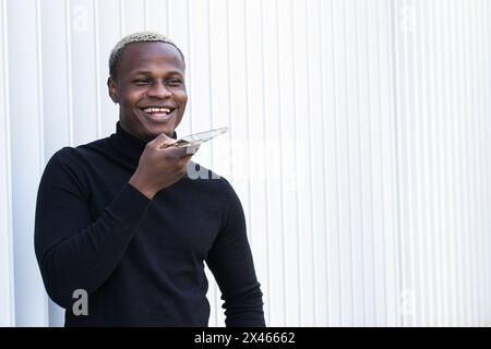 Cheerful young African American guy in black turtleneck smiling while talking on mobile phone using voice assistant on street Stock Photo