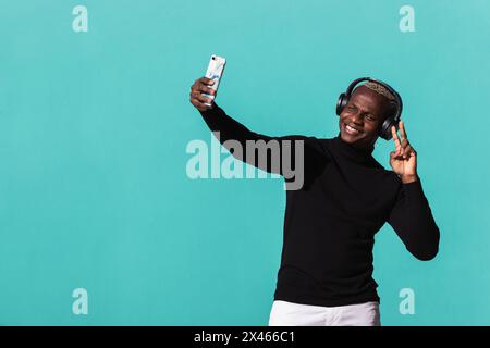 Delighted young modern ethnic guy in black turtleneck and headphones smiling and showing two fingers gesture while taking selfie on smartphone against Stock Photo