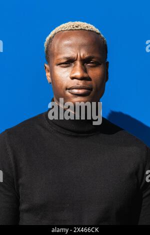 Calm young African American man with blond hair in black turtleneck standing against bright blue background on sunny day and looking at camera pensive Stock Photo