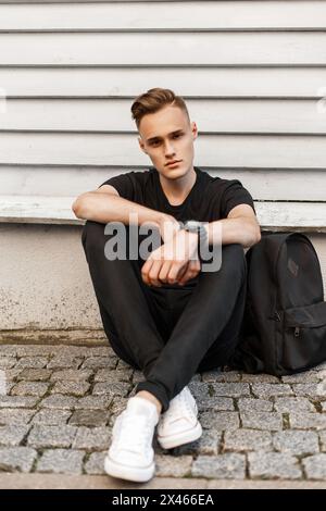 Handsome Hipster Man In A Black Shirt, Black Jeans And White Shoes With A Backpack Sitting Near The Wall. Stock Photo