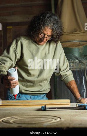 Focused mature male carpenter applying glue on piece of wood while working at table in joinery workshop Stock Photo