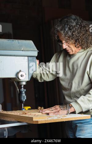 Focused mature male carpenter without a thumb drilling hole in wooden plank while working in dusty carpentry workshop Stock Photo