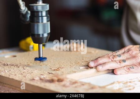 Cropped unrecognizable male carpenter without a thumb with hand tattoo drilling hole in wooden plank while working in dusty carpentry workshop Stock Photo