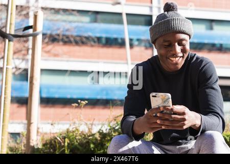 From below of African American male wearing hat and casual clothes browsing mobile phone in sunny park Stock Photo