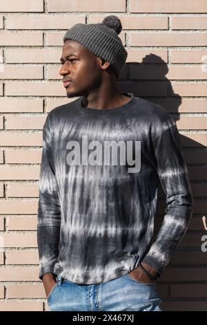 Young contemplative African American male in knitted hat standing with hands in pockets while looking away in sunlight Stock Photo