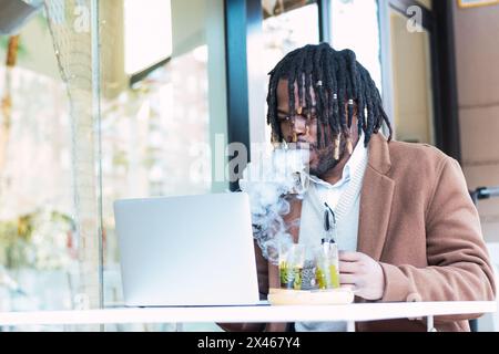 Focused adult hipster African American businessman with dreadlocks smoking cigarette while working with laptop at table with drinks in outdoor restaur Stock Photo