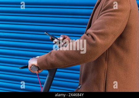 Cropped side view of unrecognizable trendy hipster African American guy in stylish coat browsing mobile phone while riding electric scooter against bl Stock Photo