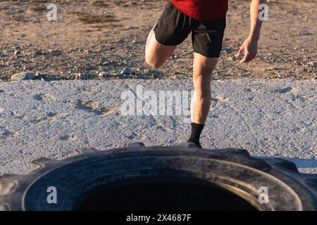 Crop anonymous male in sportswear doing stretching exercise for legs while warming up near tire on sports ground during outdoor workout Stock Photo
