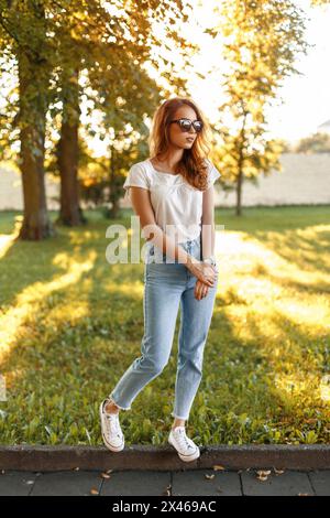 Stylish Pretty Girl In High-waisted Jeans, White T-shirt And Sneakers In A Park On A Background Of Trees At Sunset. Stock Photo