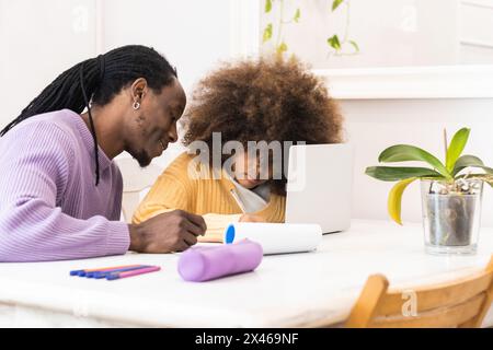 Smiling African American dad sitting at table with girl surfing netbook while preparing task for school Stock Photo