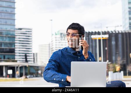 Adult ethnic cheerful male remote employee having a conversation on smartphone while working on netbook in town on sunny day Stock Photo