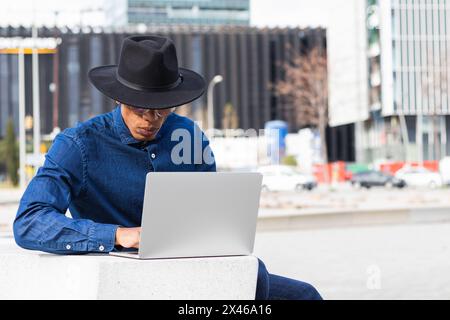 Stylish unrecognizable concentrated ethnic male remote employee in hat working on netbook in town on sunny day Stock Photo