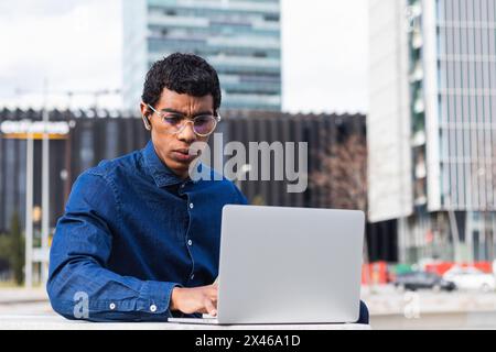 Adult concentrated ethnic male remote employee in earbud working on netbook in town on sunny day Stock Photo