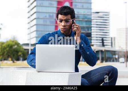 Adult concentrated ethnic male remote employee having a conversation on smartphone while working on netbook in town on sunny day Stock Photo
