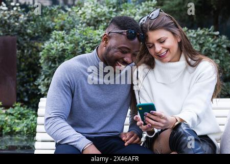 Crop view of content adult multiracial couple in love listening music on mobile phone while spending time together in park Stock Photo