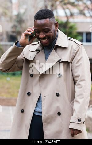 Happy adult African American male in stylish coat talking with mobile phone in hands looking away while standing on urban street in spring day Stock Photo