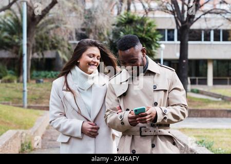 Content adult multiracial couple in love in stylish coats reading good news on mobile phone while spending time together in leafless park Stock Photo