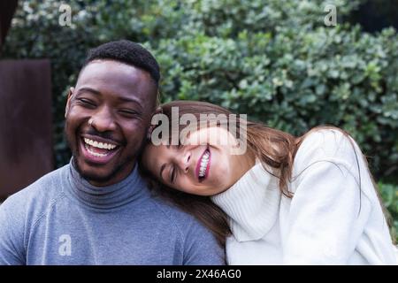 Happy young woman leaning head on shoulder of laughing African American boyfriend during romantic date in green park Stock Photo