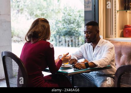 Multiethnic couple having serious conversation while sitting at table near window and having breakfast together in stylish cozy cafe Stock Photo