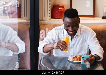 Happy adult African American male browsing mobile phone while having breakfast with glass of juice and pastry in cozy cafe Stock Photo