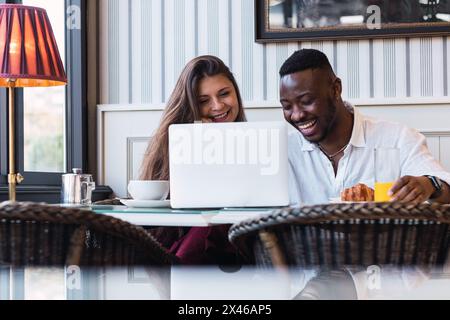 Happy multiracial couple sitting at table and browsing laptop together while having breakfast in stylish room at home Stock Photo