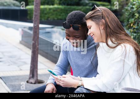 Side view of content adult multiracial couple in love reading good news on mobile phone while spending time together in park Stock Photo