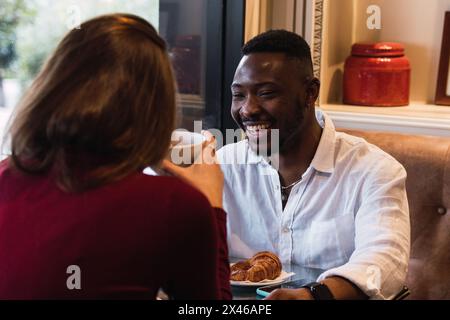 Multiethnic couple having happy conversation while sitting at table near window and having breakfast together in stylish cozy cafe Stock Photo