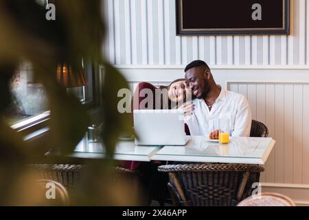 Happy multiracial couple browsing laptop together while sitting at table in stylish room at home Stock Photo