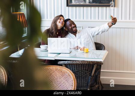 Happy multiracial couple taking a selfie while using a laptop together while sitting at table in stylish room at home Stock Photo