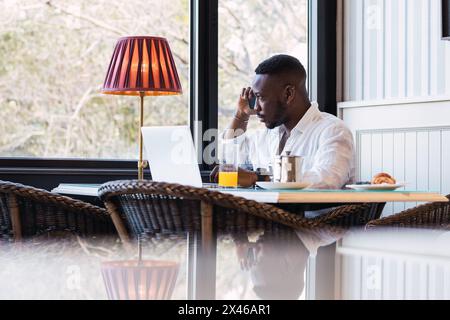 Serious adult African American businessman sitting at table with laptop and discussing business issue during phone call while having breakfast in cozy Stock Photo