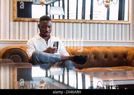 Serious young African American male in classy outfit sitting on sofa and reading news on mobile phone while resting in stylish living room with elegan Stock Photo