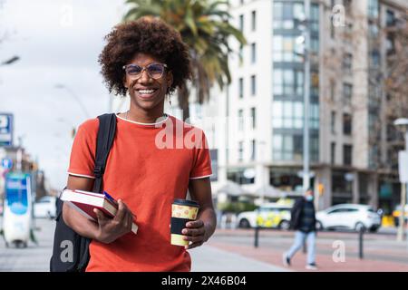 Happy young African American male student in casual clothes and eyeglasses walking on city street with books and takeaway coffee in hands Stock Photo